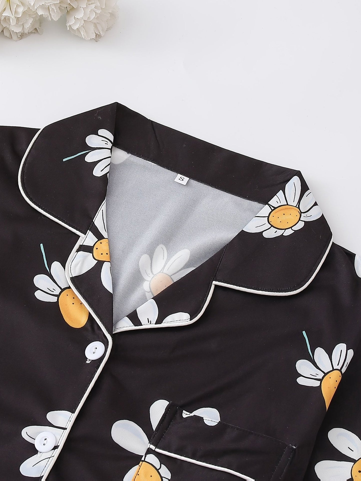 Black satin-look short-sleeve pajama set with white daisy print, piped trim, chest pocket and elastic waist shorts in 95% Polyester 5% Elastane — silky stretch loungewear