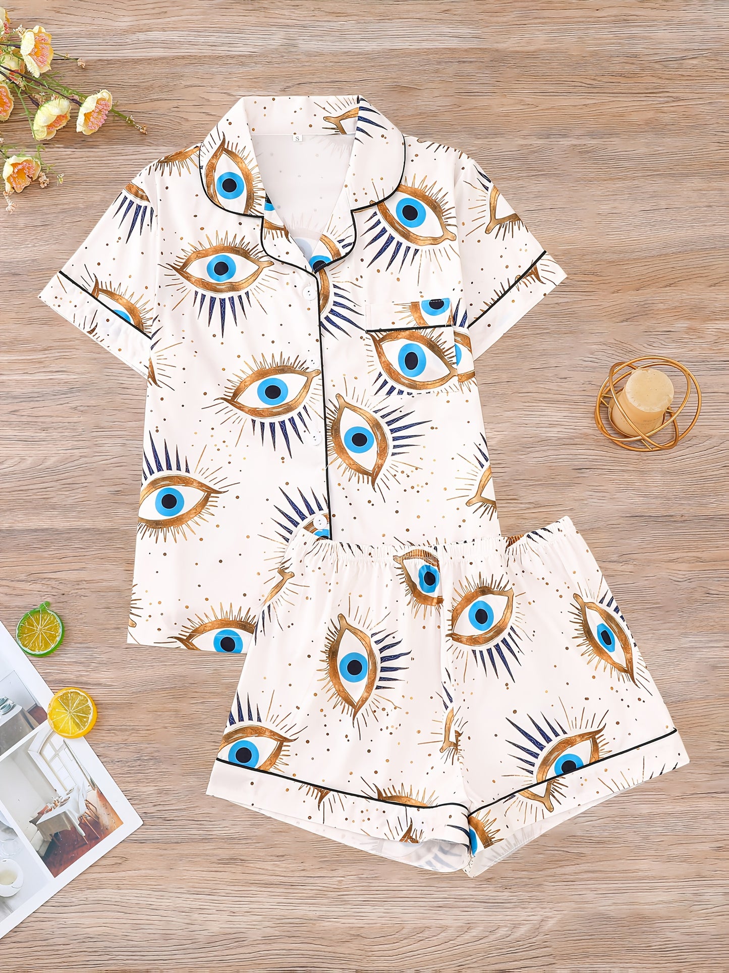 Cream satin-look pajama set with blue and gold guardian-eye print — short-sleeve button-up top with chest pocket and elastic waist shorts, 95% Polyester/5% Elastane