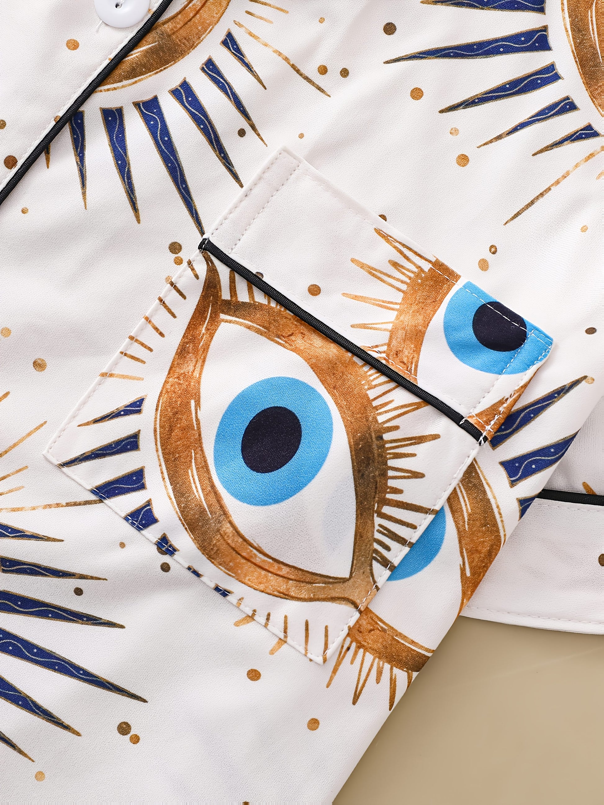 Cream satin-look pajama set with blue and gold guardian-eye print — short-sleeve button-up top with chest pocket and elastic waist shorts, 95% Polyester/5% Elastane