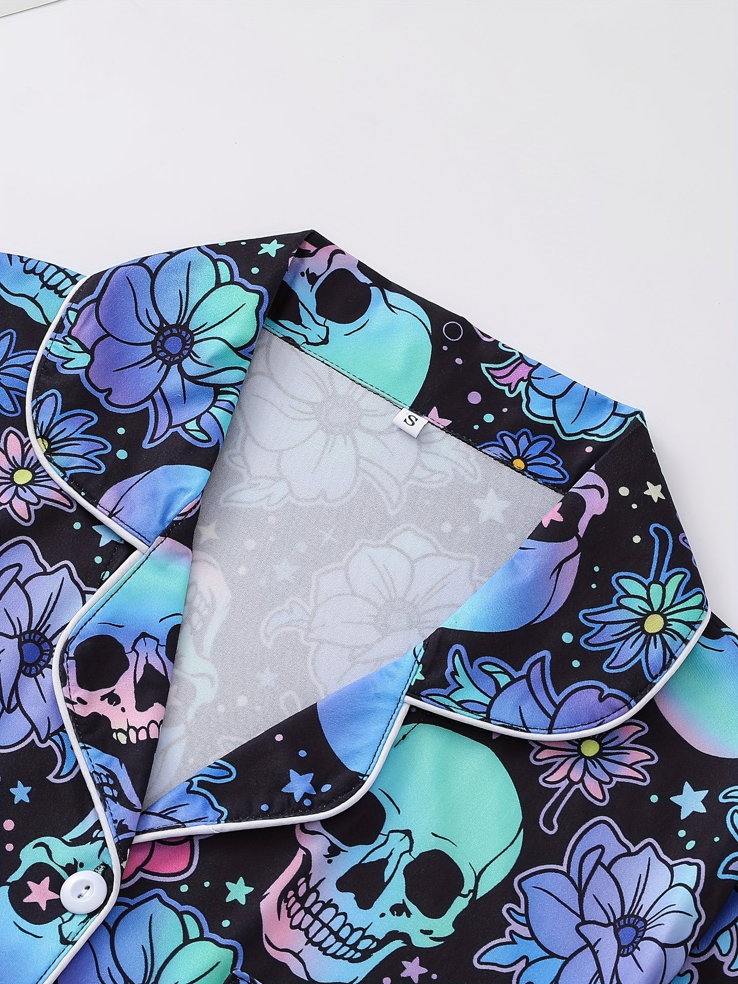 Black satin-look pajama set with neon blue and violet skull and floral print — short-sleeve button-up top with chest pocket and elastic waist shorts, 95% Polyester/5% Elastane