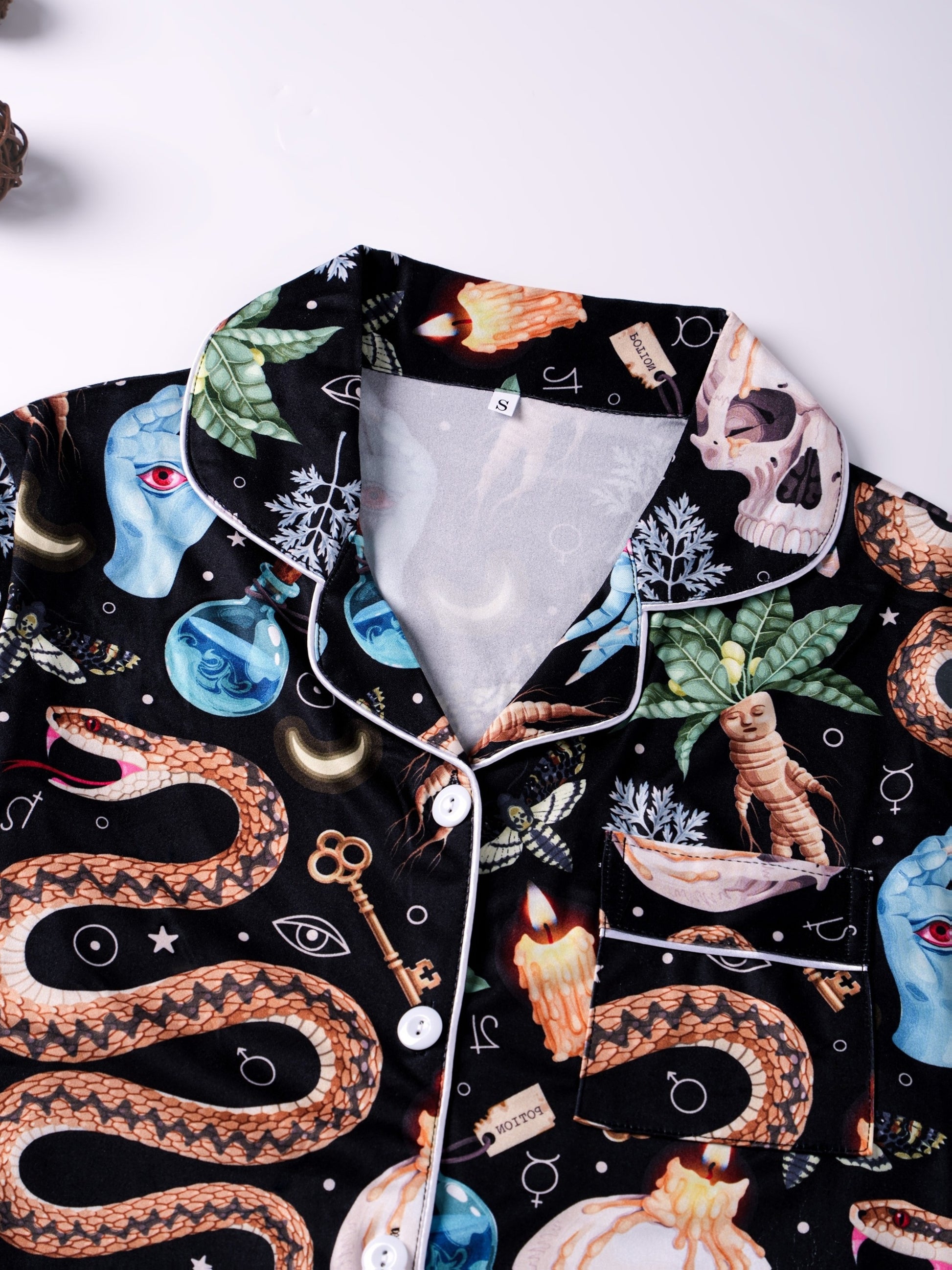 Black satin‑look pajama set with skulls, serpents, potion vials and botanicals — short‑sleeve button‑up top with chest pocket and elastic waist shorts with white piping