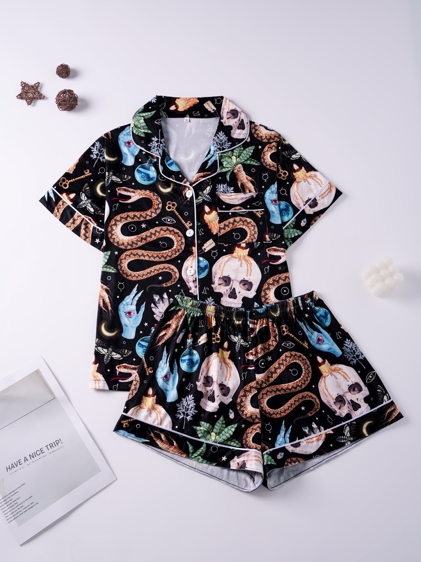 Black satin‑look pajama set with skulls, serpents, potion vials and botanicals — short‑sleeve button‑up top with chest pocket and elastic waist shorts with white piping