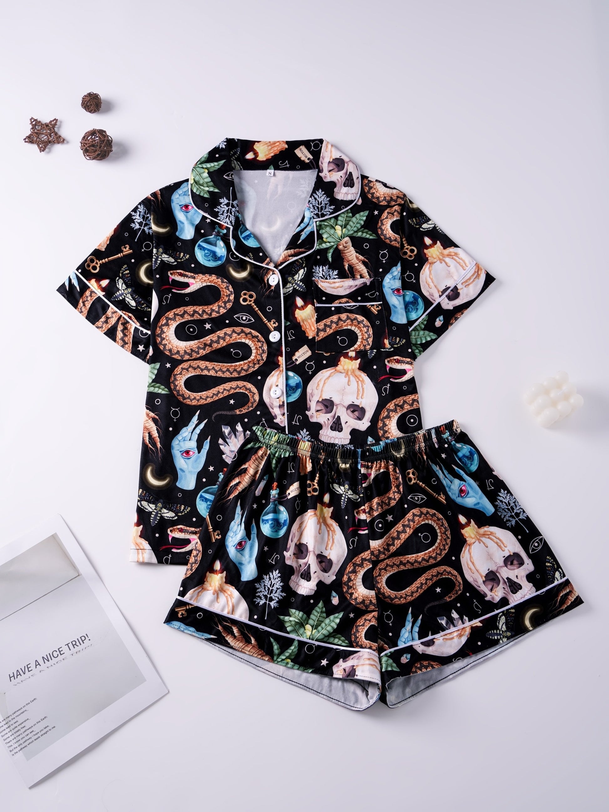 Black satin‑look pajama set with skulls, serpents, potion vials and botanicals — short‑sleeve button‑up top with chest pocket and elastic waist shorts with white piping