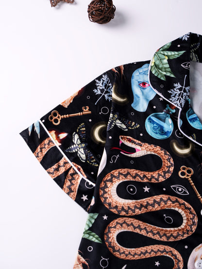 Black satin‑look pajama set with skulls, serpents, potion vials and botanicals — short‑sleeve button‑up top with chest pocket and elastic waist shorts with white piping