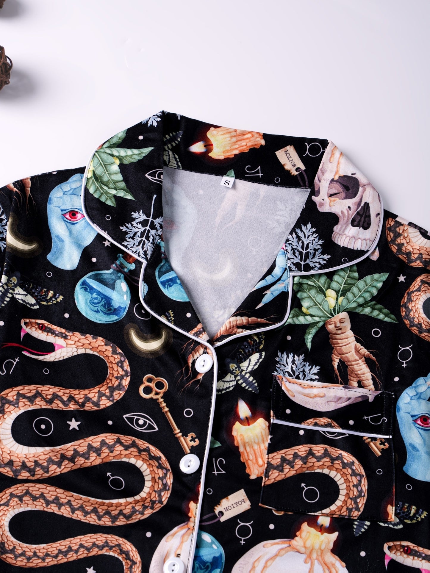 Black satin‑look pajama set with skulls, serpents, potion vials and botanicals — short‑sleeve button‑up top with chest pocket and elastic waist shorts with white piping