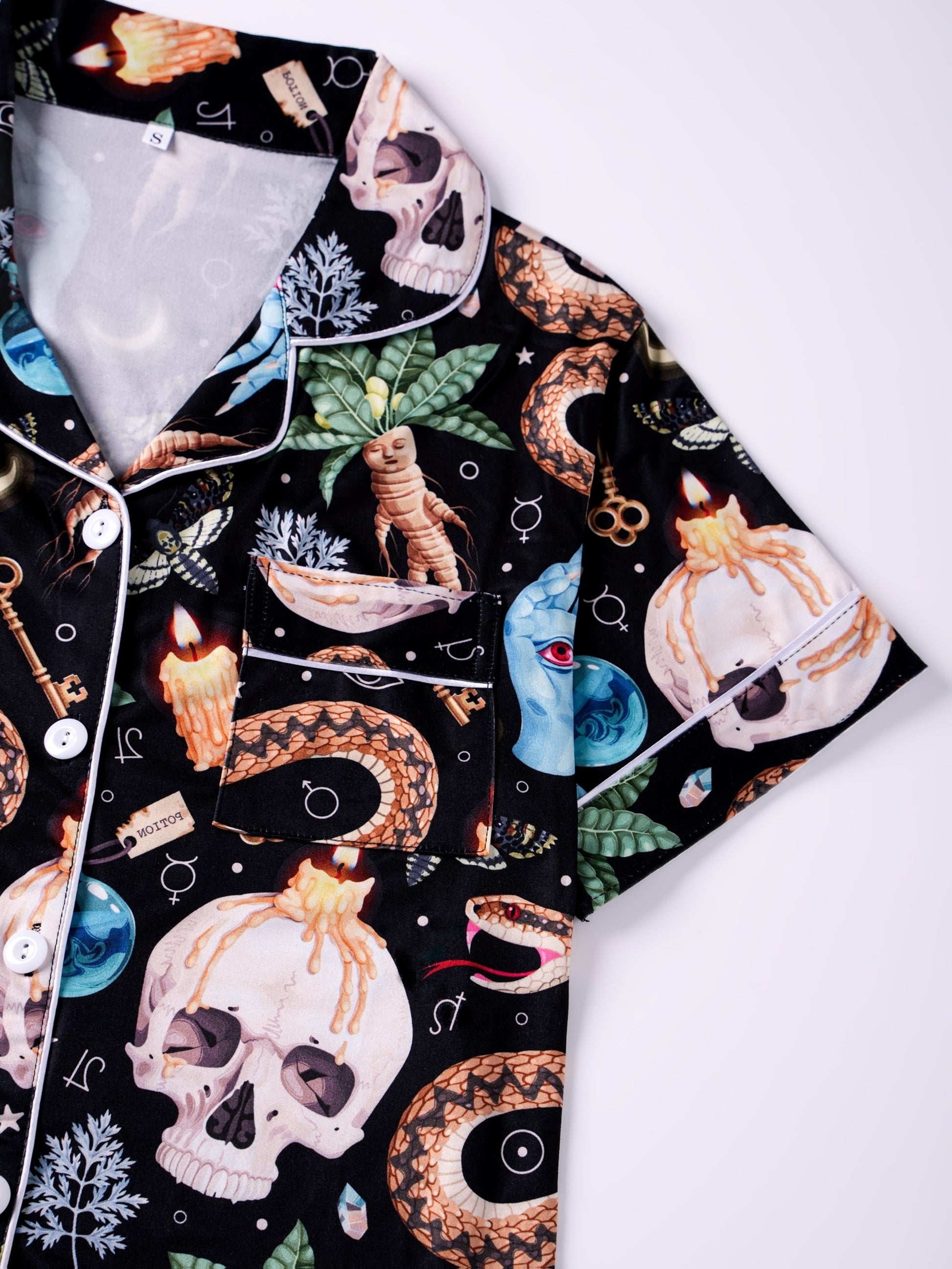 Black satin‑look pajama set with skulls, serpents, potion vials and botanicals — short‑sleeve button‑up top with chest pocket and elastic waist shorts with white piping