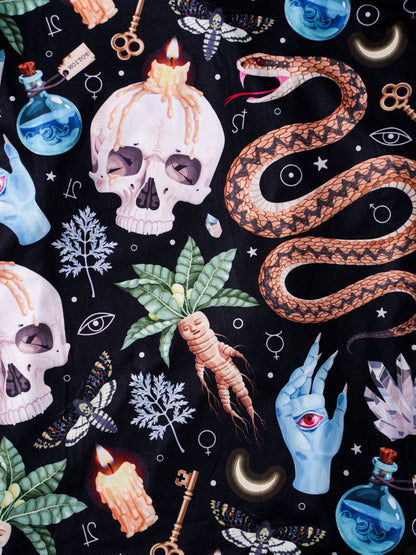 Black satin‑look pajama set with skulls, serpents, potion vials and botanicals — short‑sleeve button‑up top with chest pocket and elastic waist shorts with white piping