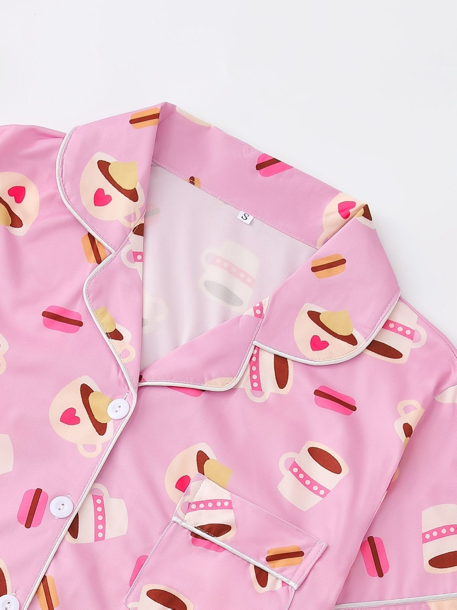 Pink satin-look short-sleeve pajama set with coffee cup and macaron print, piped trim, chest pocket and elastic waist shorts in 95% Polyester 5% Elastane — silky stretch travel-ready loungewear