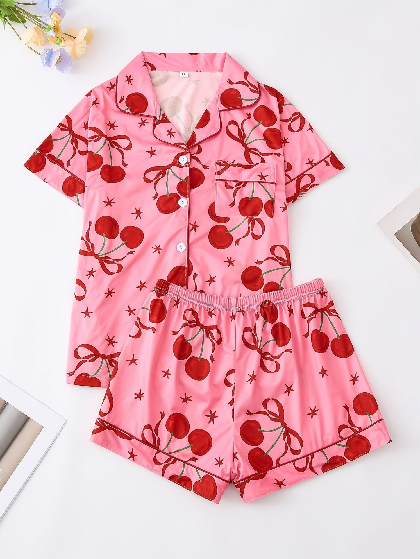 Pink satin-look short-sleeve pajama set with red cherry and bow print, piped trim, chest pocket and elastic waist shorts in 95% Polyester 5% Elastane