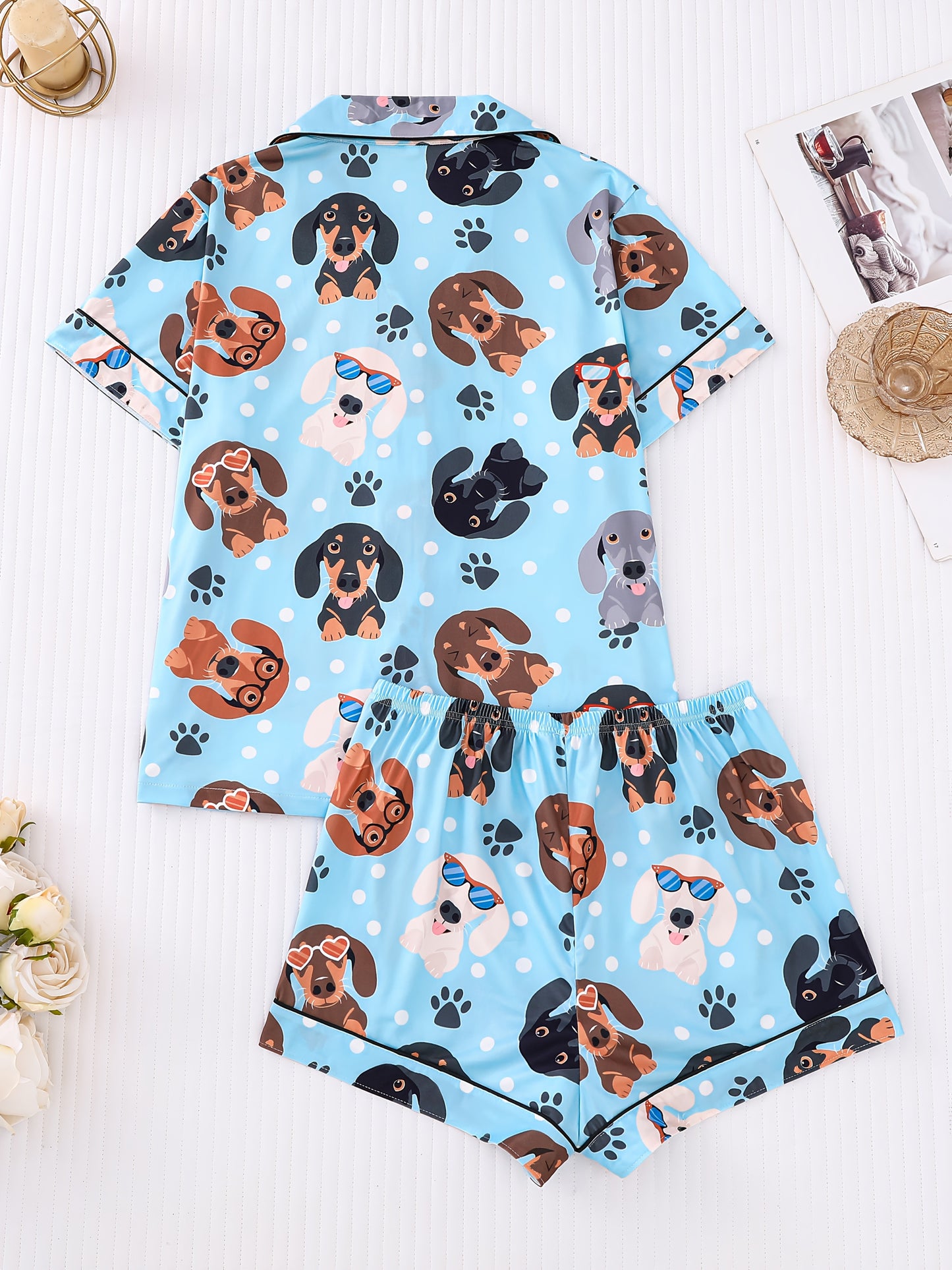 Sky-blue satin-look pajama set with playful dachshund and paw-print pattern — short-sleeve button-up top with chest pocket and elastic waist shorts, 95% Polyester/5% Elastane