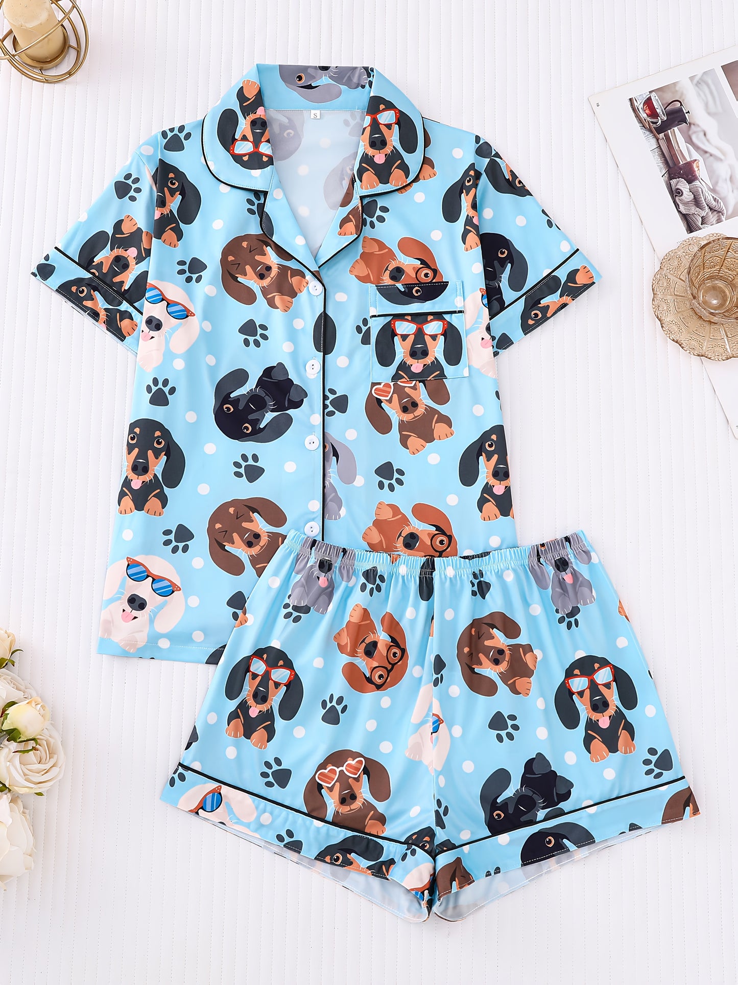Sky-blue satin-look pajama set with playful dachshund and paw-print pattern — short-sleeve button-up top with chest pocket and elastic waist shorts, 95% Polyester/5% Elastane