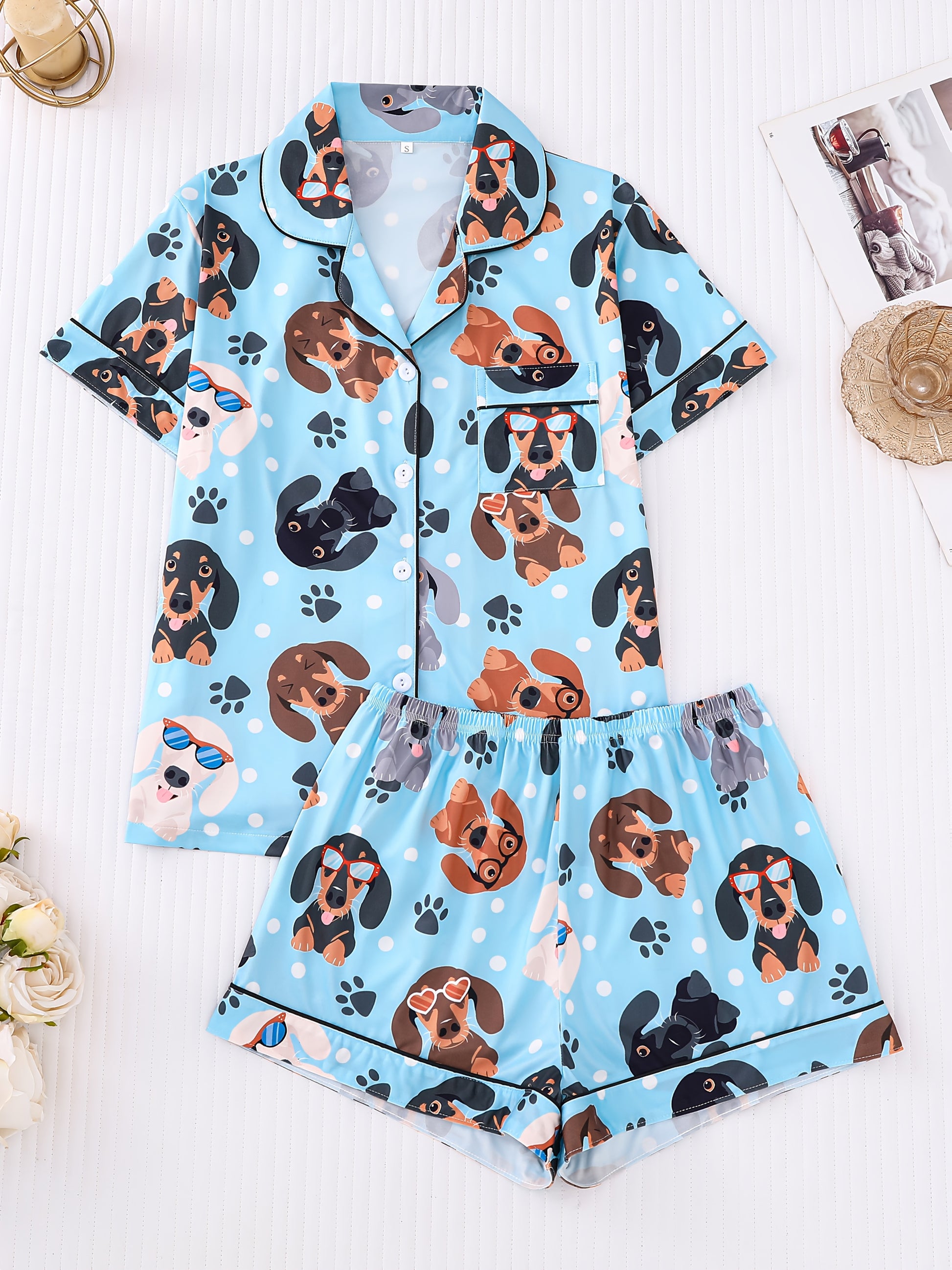 Sky-blue satin-look pajama set with playful dachshund and paw-print pattern — short-sleeve button-up top with chest pocket and elastic waist shorts, 95% Polyester/5% Elastane