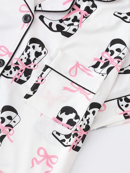 White cow-print short-sleeve pajama set with pink bow accents in silky 95% Polyester 5% Elastane, featuring chest pocket and elastic waist shorts