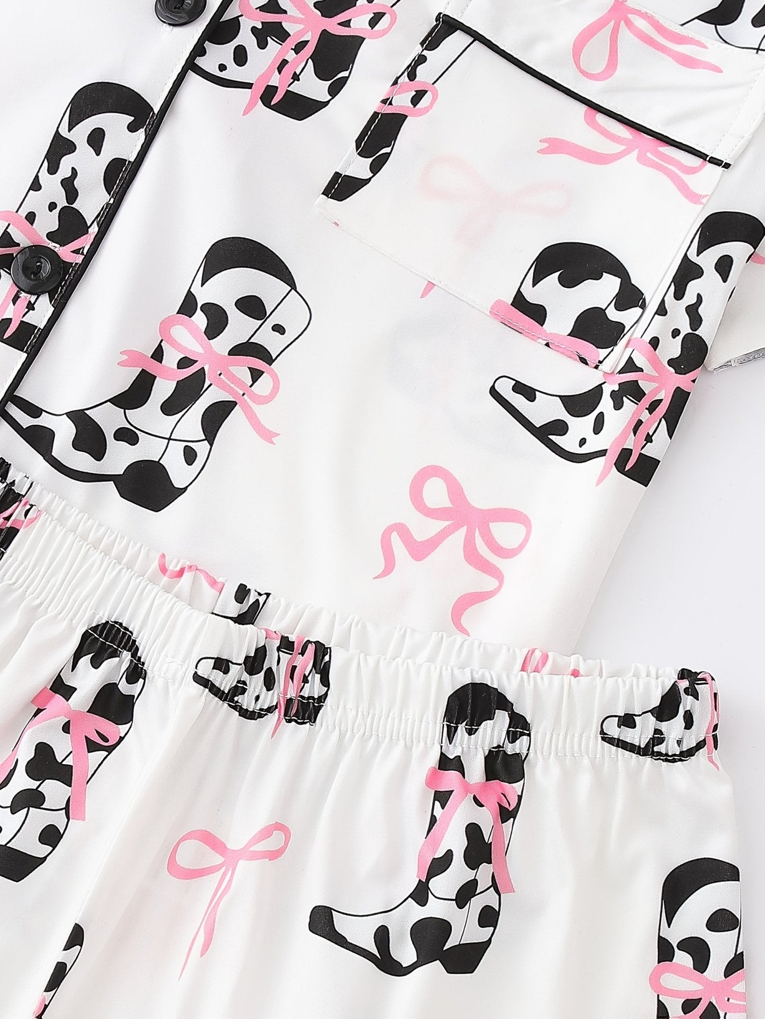 White cow-print short-sleeve pajama set with pink bow accents in silky 95% Polyester 5% Elastane, featuring chest pocket and elastic waist shorts