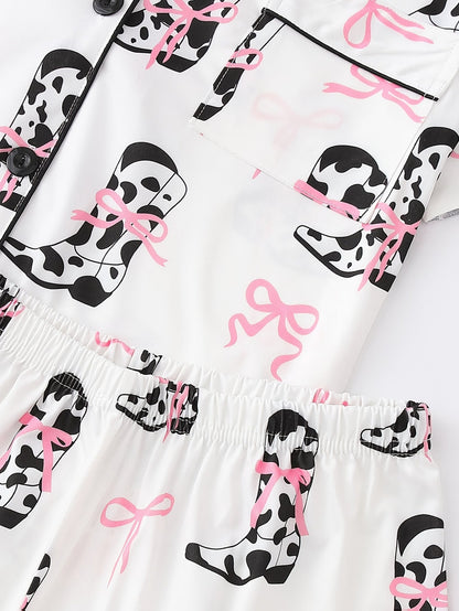White cow-print short-sleeve pajama set with pink bow accents in silky 95% Polyester 5% Elastane, featuring chest pocket and elastic waist shorts